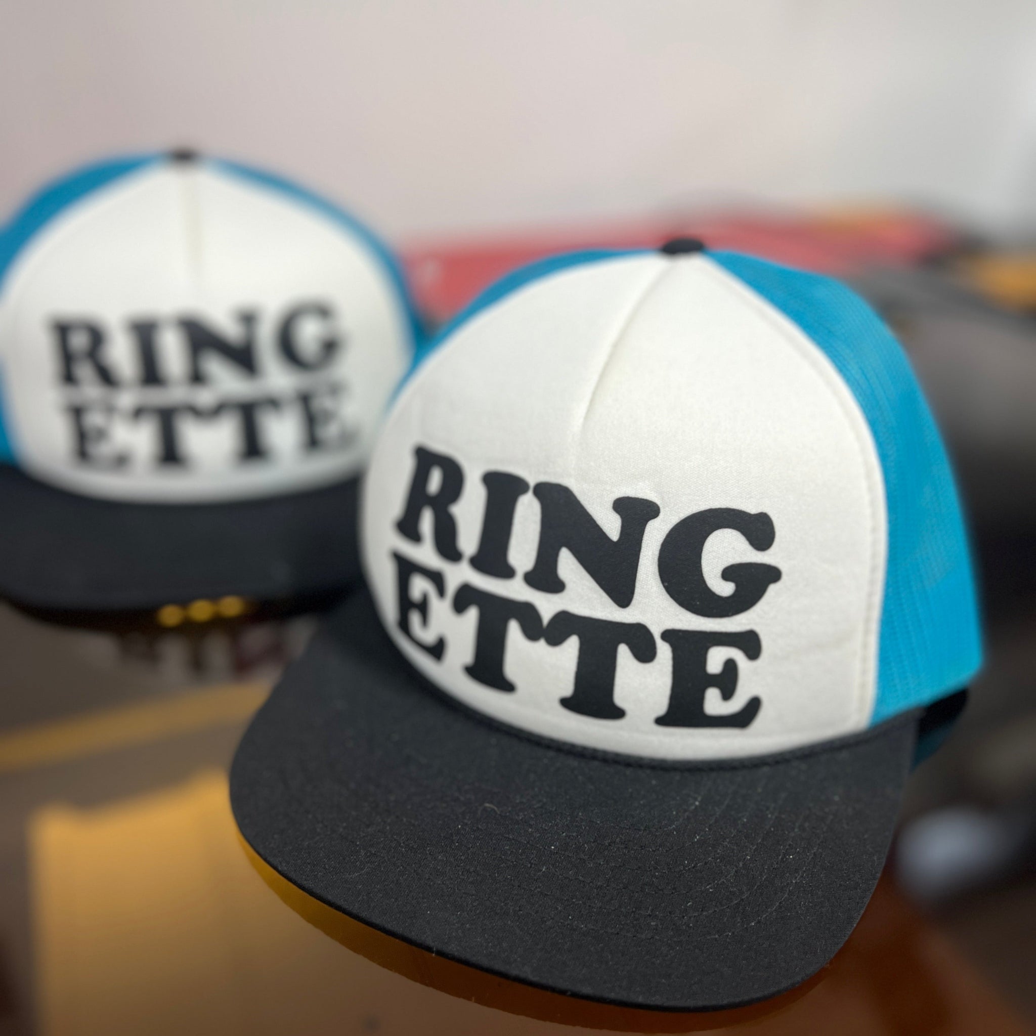 Two 'RING ETTE' caps with blue and black designs on a reflective surface.