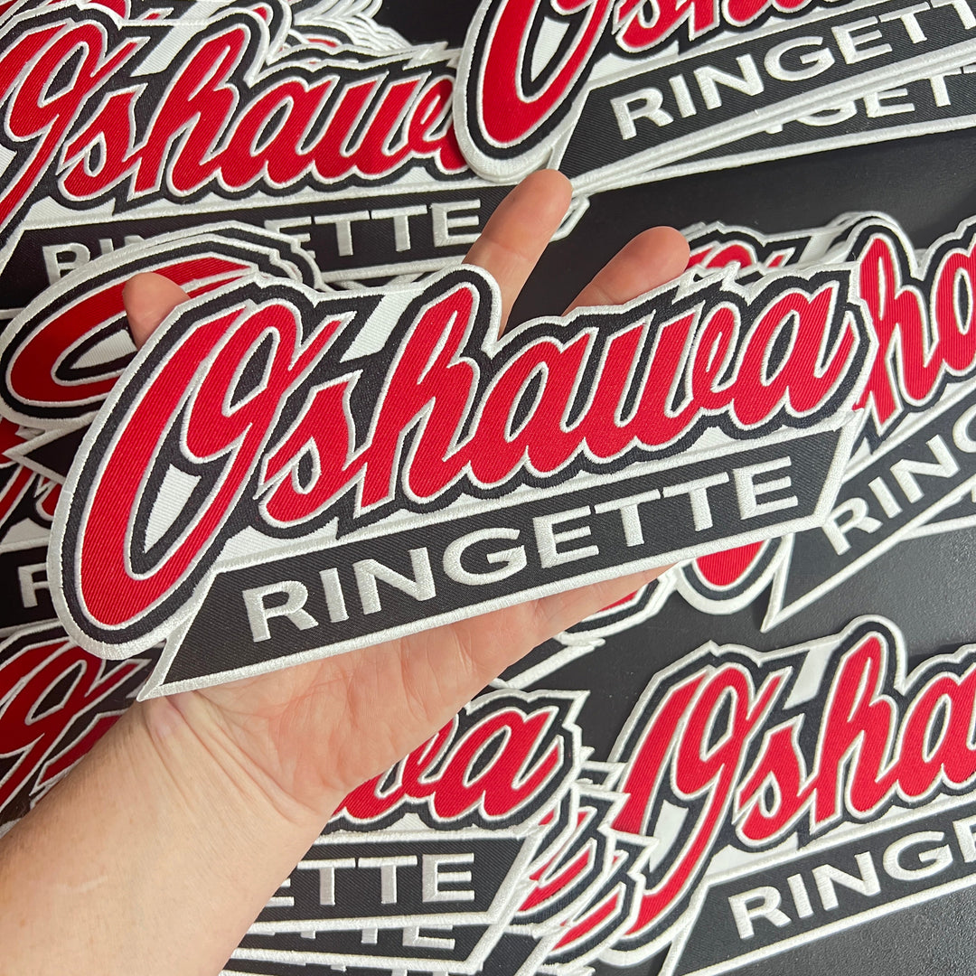 A person holding multiple oversized patches with 'Oshawa Ringette' text in red and white colors on a black background.